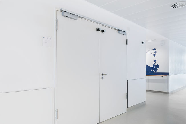 Fire protection door with TS 5000 R-ISM and hold-open function via hold-open magnet, installed at the Olgahospital in Stuttgart. Fire section door with TS 5000 R-ISM, overhead door closer with guide rail for double leaf doors with closing sequence control and smoke switch, hold-open function via hold-open magnet.