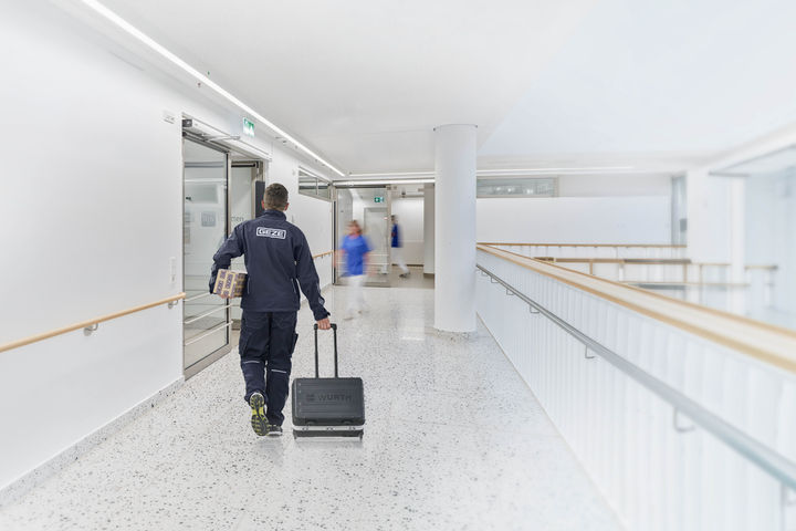 Ein GEZE-Servicetechniker läuft mit Werkzeugkoffer und Türschließer durch einen hellen Krankenhausflur