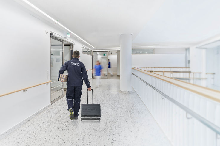 GEZE Servicetechniker läuft mit Werkzeugkoffer und Türschließer einen Krankenhausflur entlang