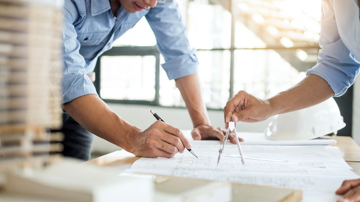 [Translate to English:] Zwei Architekten besprechen Pläne eines Gebäudes in einem modernen, lichtdurchfluteten Büro