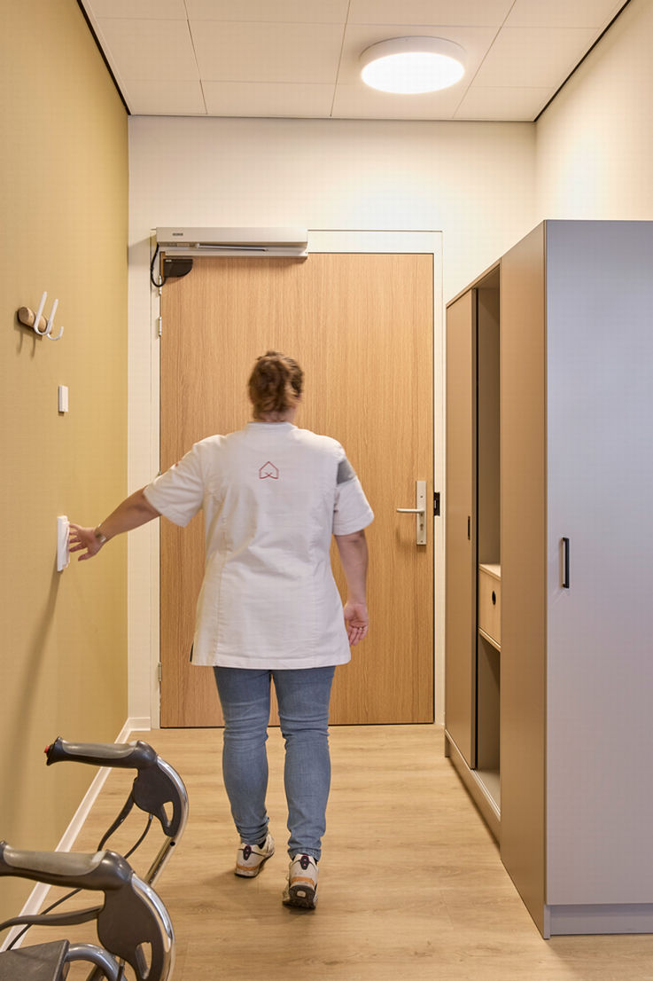 A nurse walks towards a door and opens it using a manual switch on the wall.