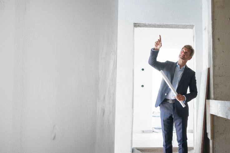 Mann im Anzug zeigt auf einen Plan in einem renovierten Raum.