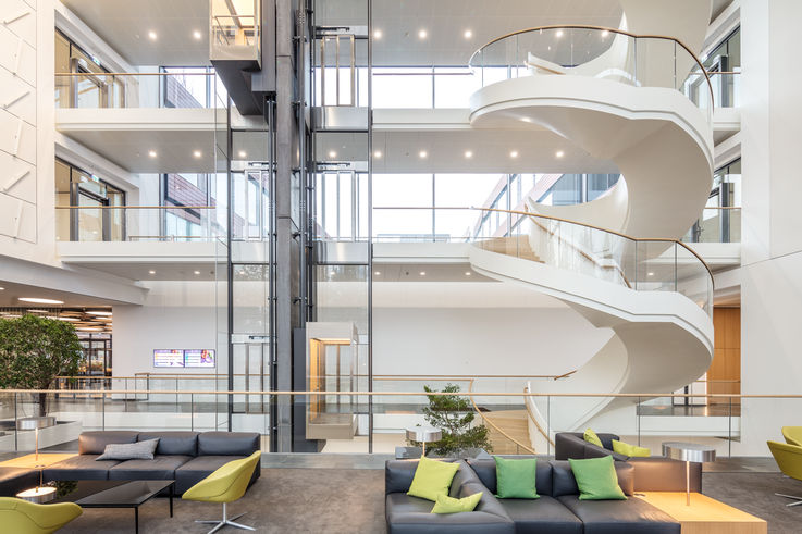 Heller, moderner Eingangsbereich eines Bürokomplexes mit großen Glasflächen, gläsernem Aufzug und Wendeltreppe.
